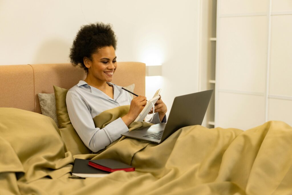 Smiling woman working remotely from bed, using laptop and taking notes.